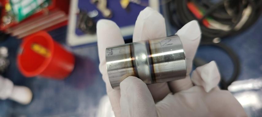 Gloved hand holding a weld coupon with gold and purple heat-tint bands along the weld seam